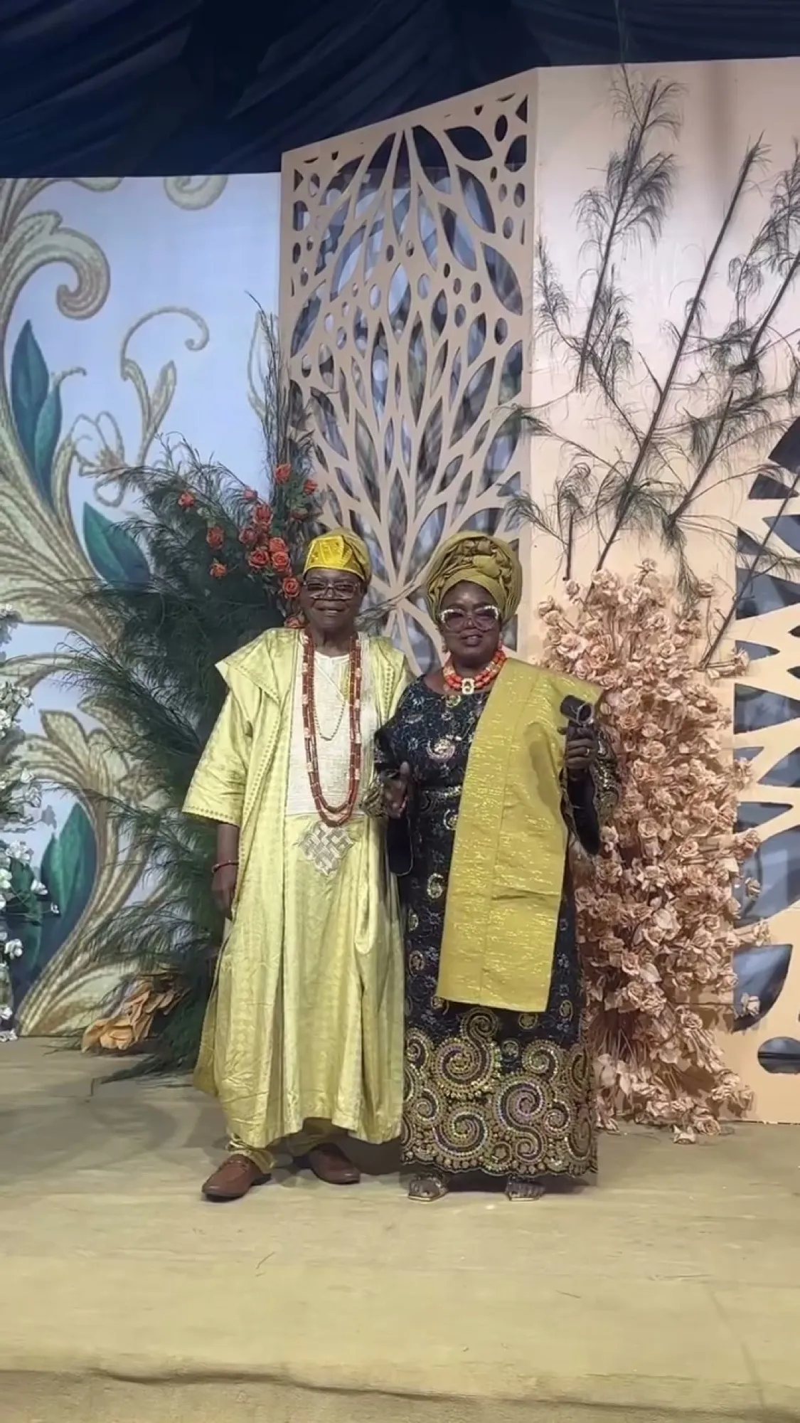 The celebrants in gold aso-oke in front of a carved floral backdrop